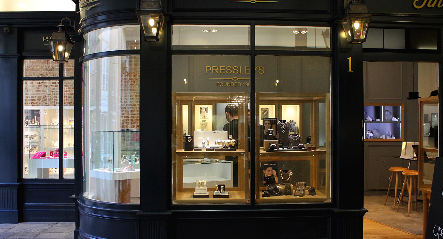 The Pressleys Jewellers shopfront in the Chichester Buttermarket at dusk, with champagne-gold lettering on the dark Regency arcade stone